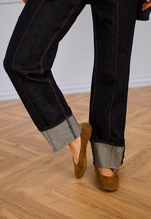 Dark denim jeans with rolled cuffs, featuring grey fabric contrast and orange stitching, paired with brown suede slip-on shoes.
