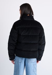 Giacca puffer in velluto a coste nera con colletto alto e design trapuntato. Presenta polsini elastici e un dettaglio logo sulla manica.