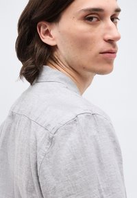 Νεαρός με μεσαίου μήκους σκούρα μαλλιά που φοράει ανοιχτό γκρι λινό πουκάμισο, κοιτάζοντας πάνω από τον ώμο του σε απλό φόντο.