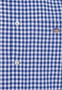 Camicia a quadri blu e bianchi con taschino e logo. Ha bottoni in plastica e una texture in cotone liscia.