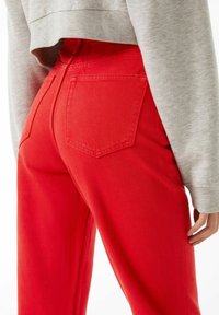 Pantalones de mezclilla rojos de cintura alta, con dos bolsillos traseros y una textura suave. Combinados con una sudadera gris corta.