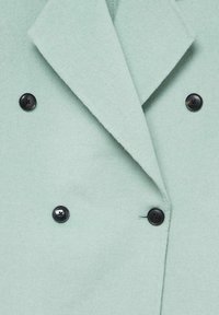 Manteau en laine vert clair doux avec un large col et quatre boutons noirs visibles disposés en deux rangées verticales.