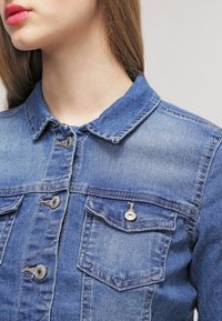 Džínová bunda s jasně modrým opráním, knoflíkovou přední částí, dvěma náprsními kapsami a klasickým límcem. Disponuje kovovými knoflíky a prošitými detaily.