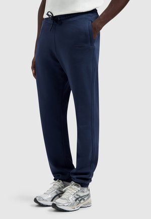 Persoon met marineblauwe joggingbroek met trekkoord en zilveren hardloopschoenen, staand met één hand in de zak tegen een effen achtergrond.