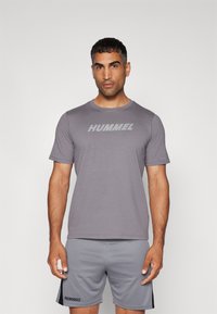 Camiseta atlética gris de manga corta y cuello redondo, que presenta un gran logotipo "HUMMEL" en el pecho. Combinada con pantalones cortos grises.