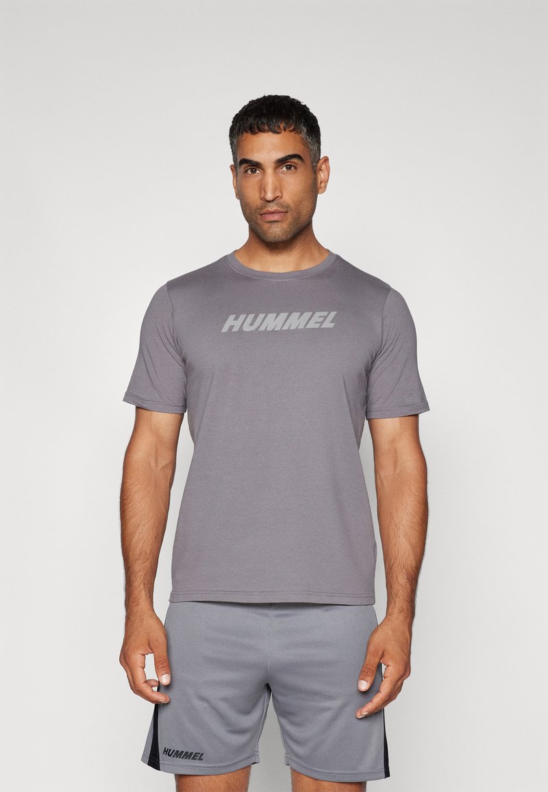 Camiseta atlética gris de manga corta y cuello redondo, que presenta un gran logotipo "HUMMEL" en el pecho. Combinada con pantalones cortos grises.