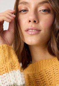 Suéter de punto con un patrón suelto y texturizado en mostaza y crema. Primer plano de rasgos faciales, piel suave y maquillaje sutil.