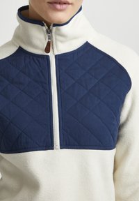 Polarni pulover s visokim ovratnikom; krema boje s mornarsko modrimi prešitimi paneli. Ima sprednji zadrgo z usnjenim ročajem.