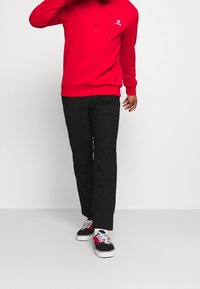 Sudadera roja con cordones y bolsillo frontal, combinada con pantalones negros. El conjunto se completa con zapatillas deportivas negras y rojas con un patrón.