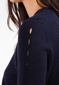 Mörkblå stickad tröja med rundad krage och sprund längs vänster ärm. Mjuk textur med en figurnära silhuett.