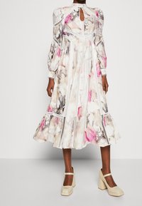 Robe à motif floral avec de longues manches bouffantes, jupe plissée, accents blancs et détail dos en goutte d'eau, complétée par des chaussures à talons blocs beiges.