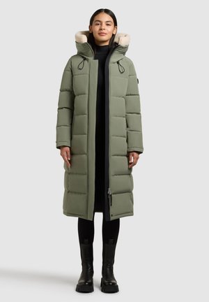 Groene gewatteerde lange jas met een capuchon van nepbont, staand model dat een zwarte coltrui en broek draagt, en zwarte imposante laarzen.