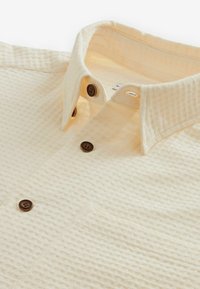 Vaffeltekstureret beige skjorte med knap-Down krave, prydet med brune knapper og en brystlomme, lavet af let stof.