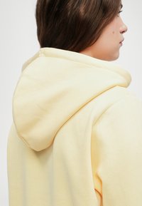 Gul hettegenser vist fra baksiden, med en myk tekstur og en avrundet hette, samt synlige sømmer nær halsen.