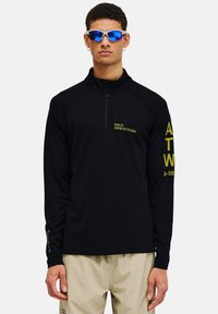 Schwarzer Langarmpullover mit Reißverschlusskragen, der gelben Text auf der Brust und dem Ärmel aufweist und aus strukturiertem Material besteht. Trägt eine Sonnenbrille.