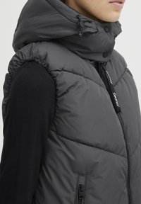 Gilet matelassé gris avec col haut et capuche réglable, doté de poches avec fermeture éclair et d'une étiquette logo sur la tirette de la fermeture. Texture lisse.