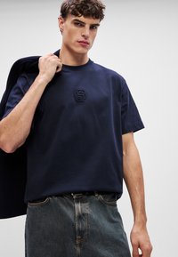 Μπλε βαμβακερό μπλουζάκι με κοντά μανίκια, διακοσμημένο με υφασμάτινο λογότυπο στο στήθος, συνδυασμένο με ξεθωριασμένα τζιν παντελόνια.