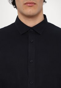 Camicia nera con colletto a punta, realizzata in un tessuto testurizzato. Presenta bottoni neri e un logo discreto sul petto.