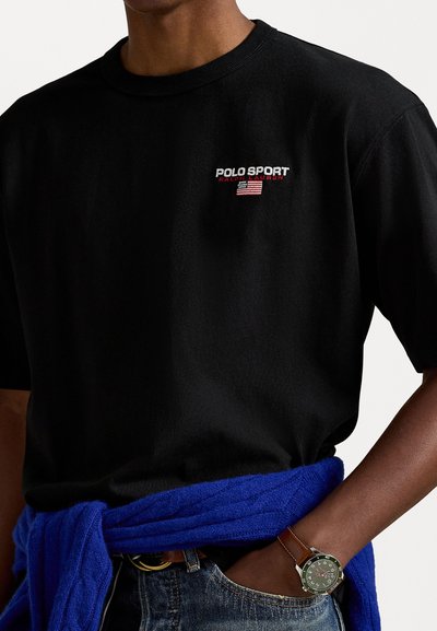 Camiseta de algodón negra con el logo de "POLO SPORT" en rojo y blanco, de mangas cortas y cuello redondo, acompañada de un suéter de punto azul atado a la cintura.