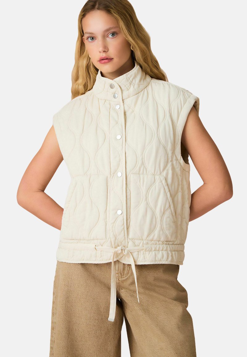 Gilet sans manches en crème matelassé avec col montant, fermetures à pression et lien ajustable à la taille ; présente une texture ondulée et des poches latérales.