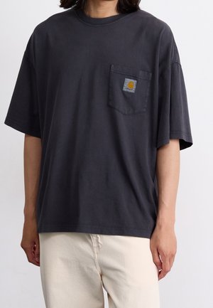 Personne portant un t-shirt Carhartt ample gris foncé avec logo sur la poche et un pantalon beige clair, debout devant un fond blanc uni.