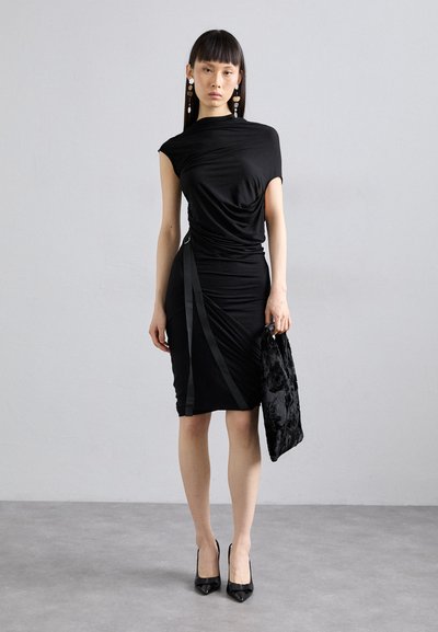 Vestido preto justo com detalhe drapeado, mangas curtas e acabamentos semelhantes a cetim. Usado com sapatos de salto alto e uma bolsa texturizada preta.