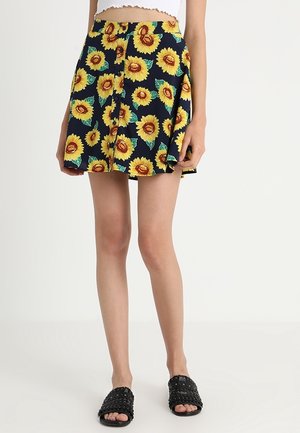 Frau trägt einen nachtblauen Rock mit großem gelbem Sonnenblumenmuster, weißes kurzes Oberteil und schwarze Sandalen mit Nieten.