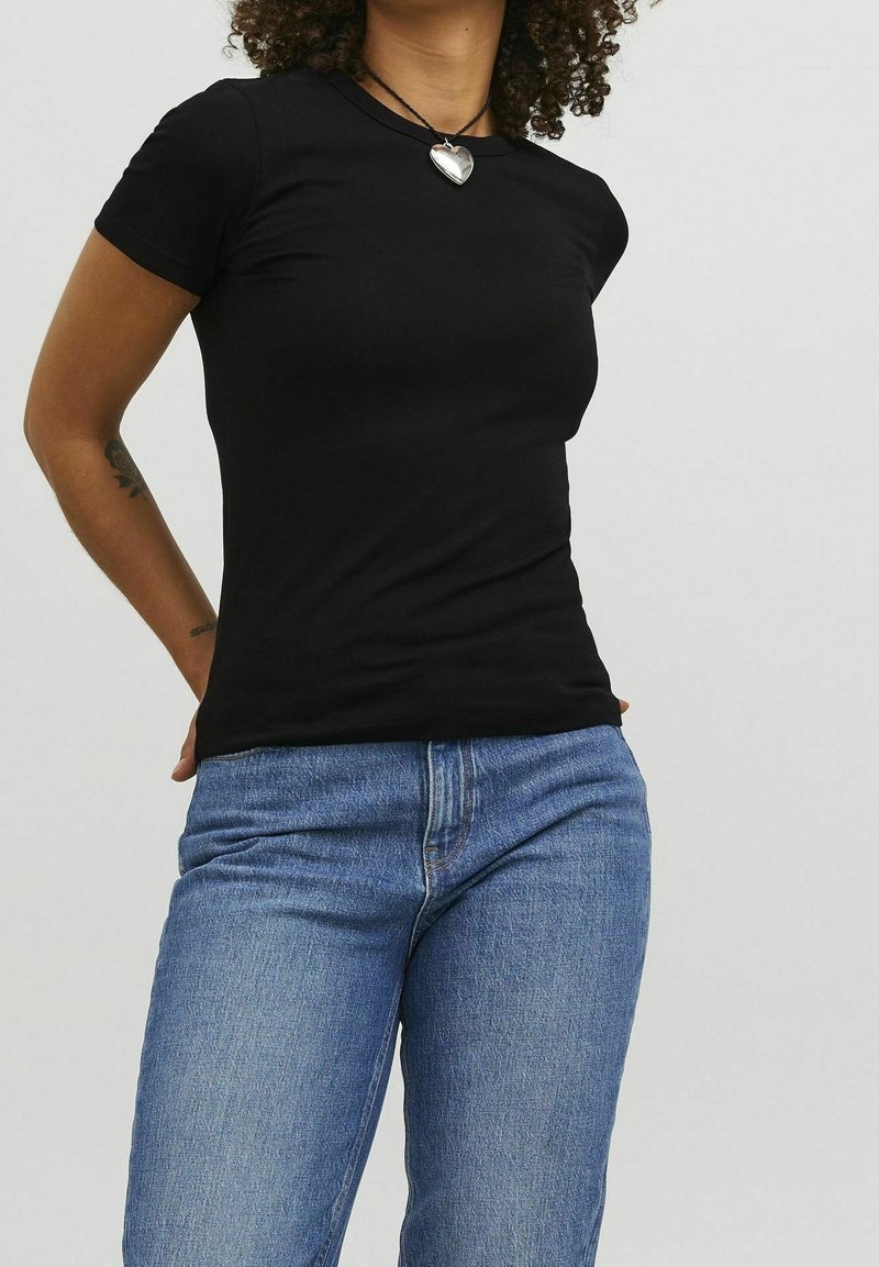 Woman wearing a fitted black short-sleeve t-shirt, blue jeans, heart-shaped silver pendant necklace, with tattoo on her left forearm.