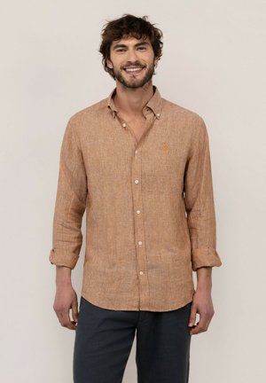 Hombre sonriente con cabello rizado lleva una camisa beige a rayas abotonada con las mangas remangadas y pantalones oscuros sobre un fondo liso y claro.