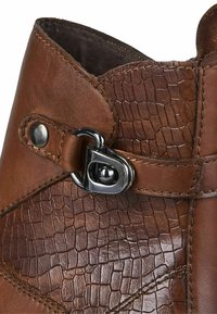 Bota de cuero marrón con un patrón texturizado que recuerda a una piel de reptil, acentuada con una hebilla metálica y un botón a presión, con un acabado suave.
