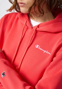Sudadera color coral con capucha ajustable, textura suave y logotipo "Champion" en blanco en el pecho. Presenta un parche con el logotipo en la manga.