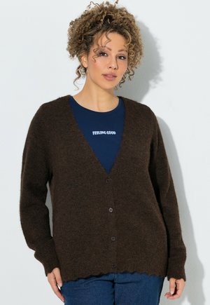 Kvinde med krøllet hår har en mørkebrun knappet cardigan over en marineblå skjorte med hvid tekst, der lyder "FEELING GOOD."