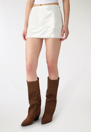 Donna con minigonna bianca di paillettes e stivali di camoscio marroni alti al ginocchio, in piedi contro uno sfondo chiaro semplice.