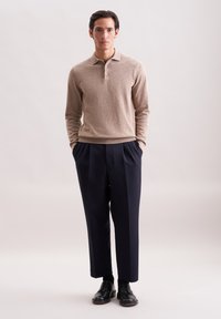 Ein Mann trägt einen beigen Langarm-Polo-Pullover, dunkle gefaltete Hosen und schwarze Anzugschuhe und steht vor einem hellen, einfarbigen Hintergrund.