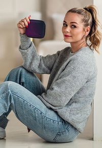 Compact black and purple wallet held by a person wearing a gray ribbed sweater and light blue jeans, sitting on the floor.