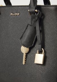 Schwarze Handtasche aus strukturiertem Material mit einem goldenen ALDO-Logo, einem Schlüssel und einem durch einen Riemen befestigten Schloss. Die Griffe sind ebenfalls schwarz.