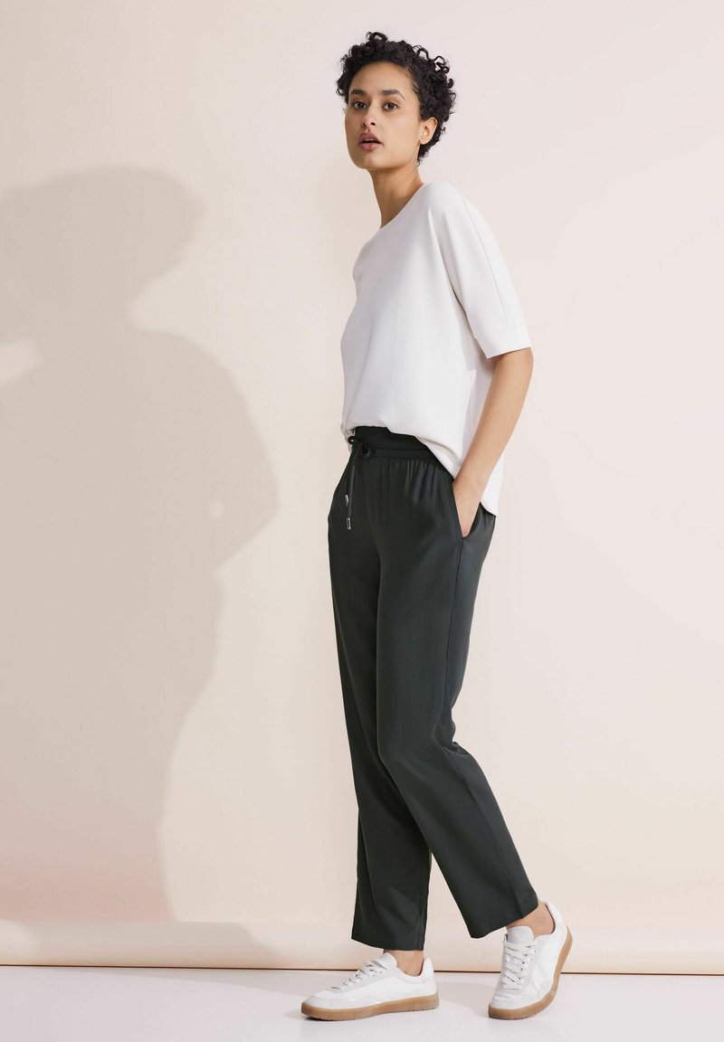 Top blanco de manga corta y pantalones verde oscuro, con cintura ajustable. Zapatillas blancas con suela marrón completan el conjunto.