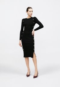 Kobieta stoi z ręką na biodrze, ubrana w czarną prążkowaną sukienkę midi z złotymi guzikami po bokach oraz bordo szpilki na obcasie z odkrytą piętą.