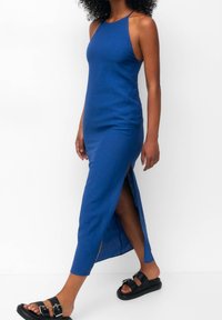 Femme portant une robe midi bleue sans manches avec une fente latérale, associée à des sandales noires à doubles brides à boucle, marchant sur fond blanc.