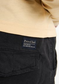 Zwarte broek met een gestructureerde afwerking, voorzien van een zichtbaar navyblauw label met de tekst "Petrol Ind" in witte letters. Een lichtgele shirt is gedeeltelijk zichtbaar.