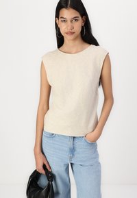 Lys beige ærmeløs top med en tekstureret overflade, rund halsudskæring og løs pasform, parret med lyseblå vidde bukser og en sort taske.