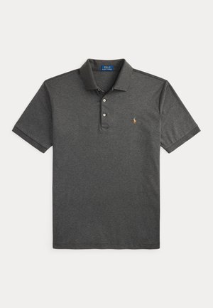 Homme portant un polo gris foncé, un jean foncé avec une ceinture marron, des mocassins marron et une montre, debout devant un fond beige uni.