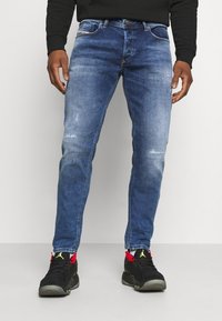 Blue denim jeans with a slim fit, featuring subtle fading and distressed details. Paired with black sneakers and a black sweatshirt.