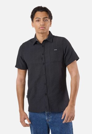 Schwarzes T-Shirt mit kurzen Ärmeln, strukturiertem Muster, Knopfleiste vorne und Brusttasche. Kombiniert mit hellblauen Jeans.