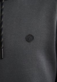 Felpa grigia scura con texture a coste, caratterizzata da un logo rotondo nero e cordini neri a contrasto. Tessuto liscio, design semplice.