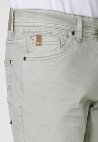 Svetlo sivi traper hlače s vidljivi prednji žep, žep za kovanice s smešnim oznakom, petlje za pojas i detajli s zakovicama.