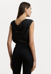 Zwarte mouwloze top met een gedrapeerde halslijn, gemaakt van een gladde stof. Gecombineerd met strakke zwarte jeans met een hoge taille en achterzakken.