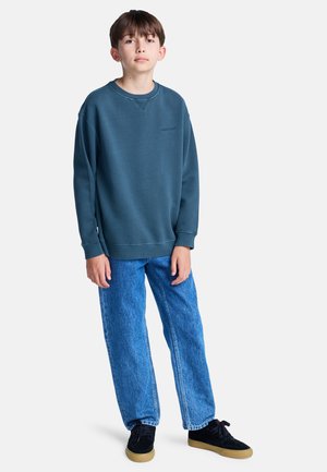 Felpa blu con scollo a girocollo e logo, abbinata a jeans dritti blu chiaro e scarpe nere con suola in gomma.