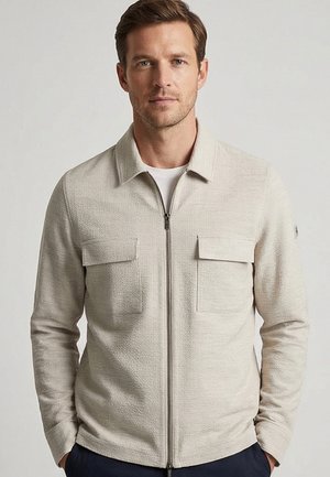 Mann trägt eine hellbeige strukturierte Reißverschlussjacke mit Kragen und zwei vorderen Klappentaschen, stehend vor hellem einfarbigem Hintergrund.