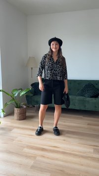 Črna bluzica s črnimi vzorci in abstraktnimi belimi črtami, kombinirana s črnimi bermudami, črnimi čevlji in črnim baretom. Vidna zelena kavča in rastlina.